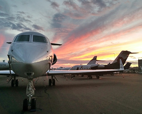 Private jets parked on an airport tarmac at sunset, with vibrant orange and pink clouds stretching across the sky behind them.