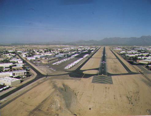 Aerial view down a long runway flanked by taxiways, hangars, aircraft parking areas, and desert terrain under a clear sky.