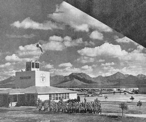 Historic-style airport scene with a building, a windsock on the roof, distant mountains, several small aircraft, and a group of people gathered on a lawn in front.
