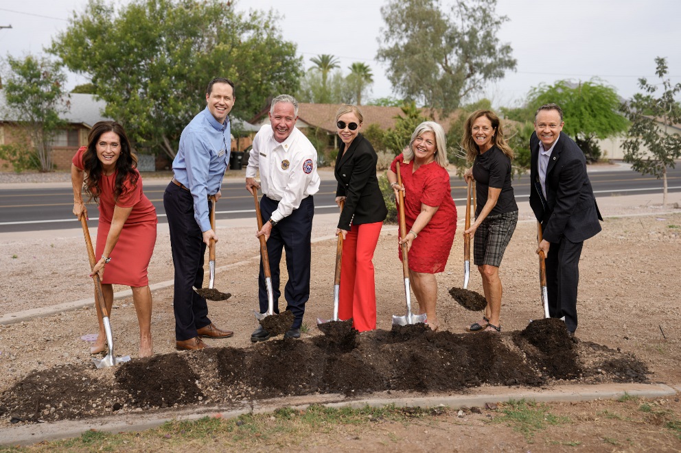 Seven people smiling during a ceremonial groundbreaking, each holding a shovel over a row of fresh soil beside a roadside construction area.