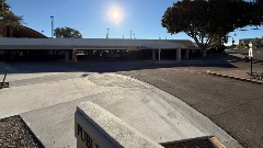Wide sidewalk and curb ramp at the entrance to a public parking garage, with vehicle driveway access, clearance signage, and bright sunlight overhead.