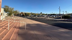 Concrete steps with handrails lead to a sidewalk beside a street and a parking lot with parked cars under clear blue skies.