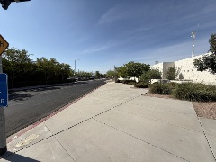 Sidewalk running alongside a landscaped area with shrubs, trees, and a modern building, bordering a freshly paved street under a clear sky