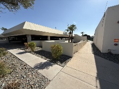 Concrete walkway bordered by low beige walls, desert landscaping, and a covered parking structure with angled rooflines under a clear sky