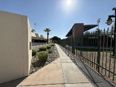 Concrete walkway beside a fence and landscaped shrubs, running between a parking structure and a stadium-style building under bright sunlight