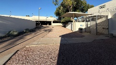 Pedestrian walkway leading toward a parking garage with adjacent City Jail entrance, fencing, signage, and shaded seating area.