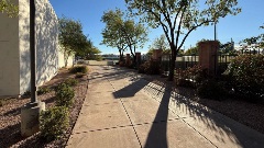 Concrete pedestrian walkway curves past a public building with desert landscaping, trees, and fencing in bright daylight.