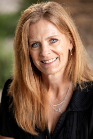 Person with long light-colored hair wearing a dark collared top, posed outdoors with a soft blurred background