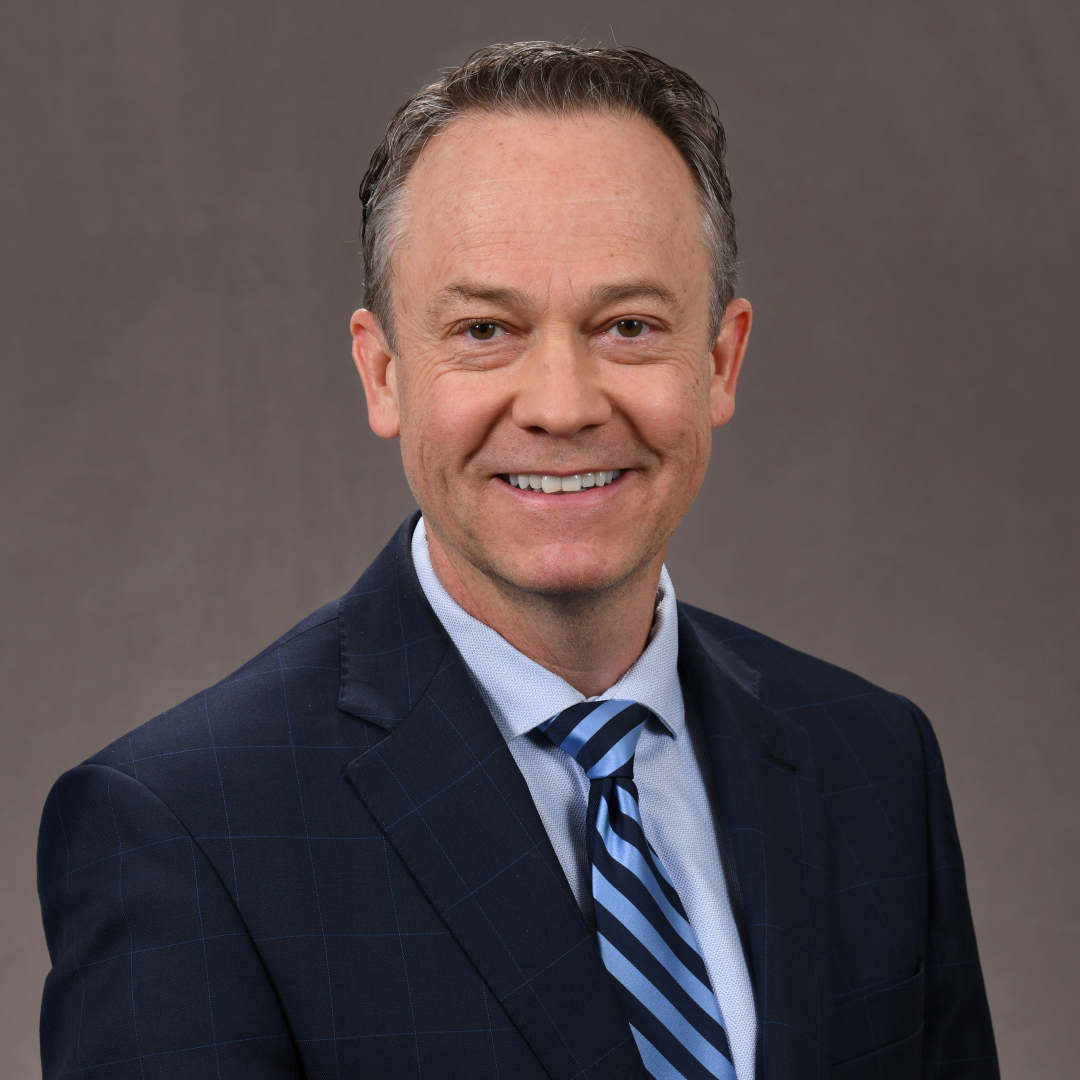 Professional headshot of a man in a navy suit and blue striped tie, smiling against a gray background.