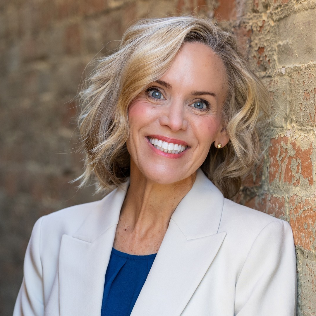 Professional headshot of a woman with blonde hair in a dark denim top, smiling in front of a light interior with a plant.