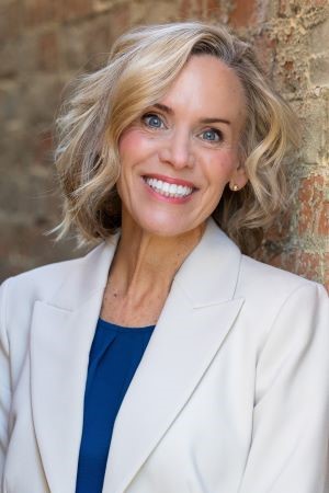 Headshot of Kira Peters smiling in a white blazer against a brick wall.