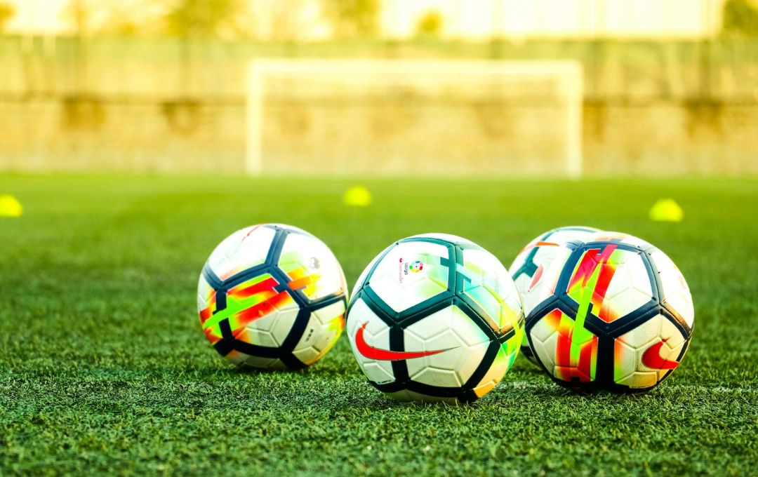 Three colorful soccer balls on a grass field with a blurred goal in the background.
