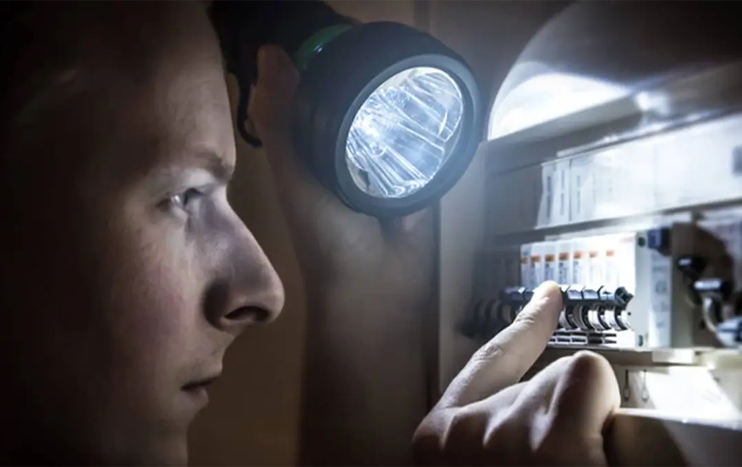 Person using a flashlight to inspect and reset switches on an electrical breaker panel.