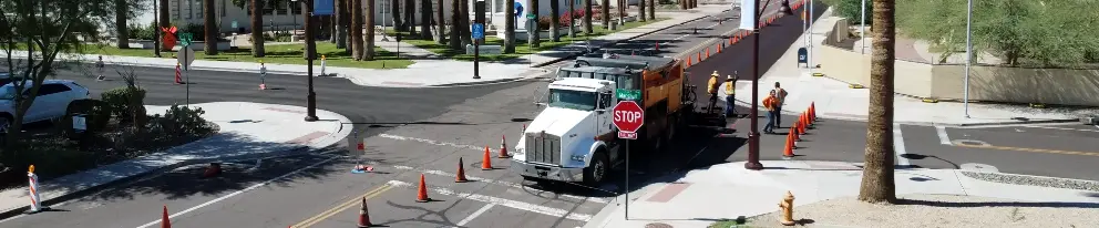 Road construction crew paving a city street intersection with a work truck, traffic cones, and workers in safety vests directing traffic near palm-lined sidewalks and crosswalks