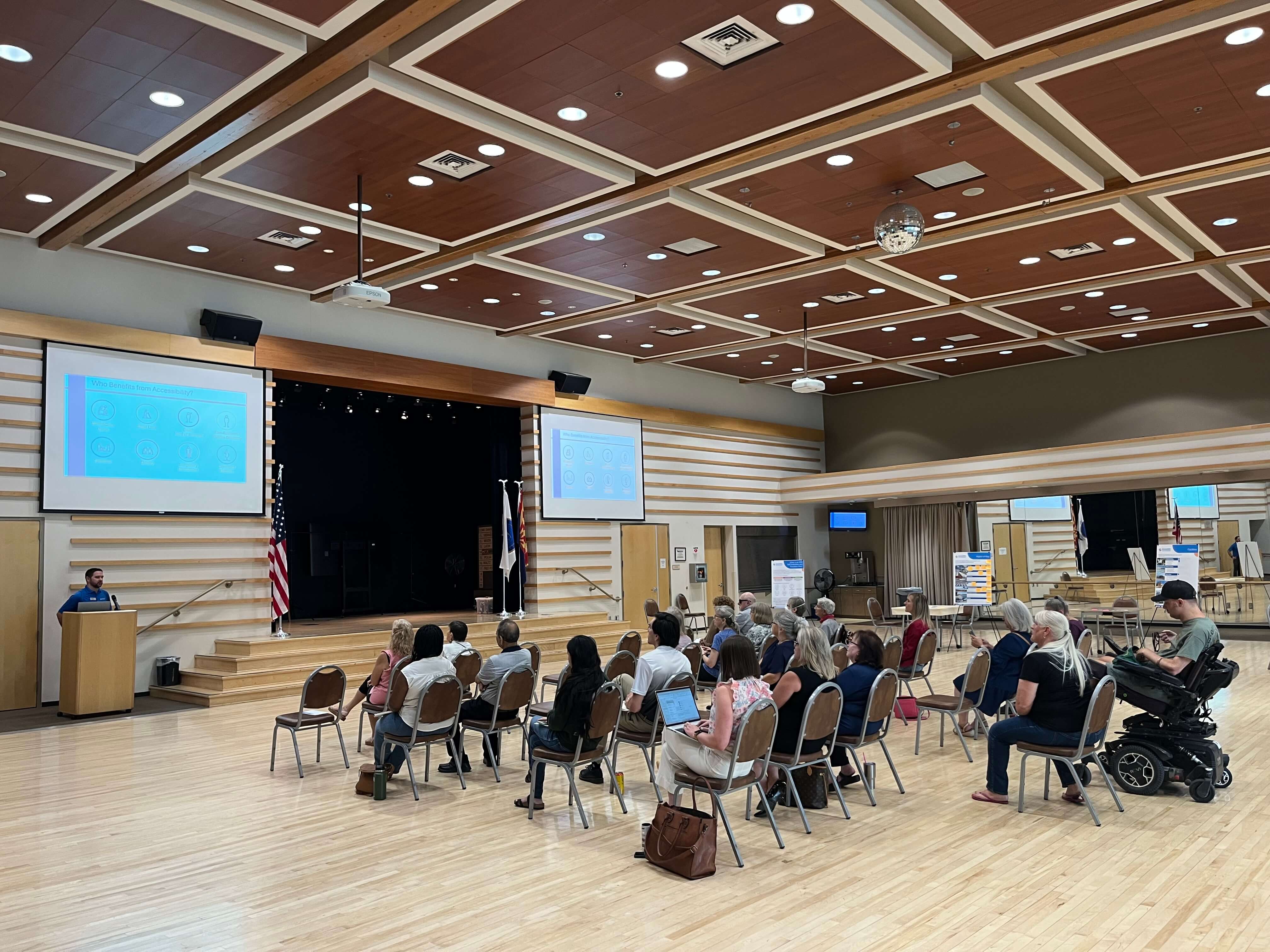 Audience seated in a community center watching a presentation about accessibility improvements, with slides displayed on large screens and informational posters around the room.