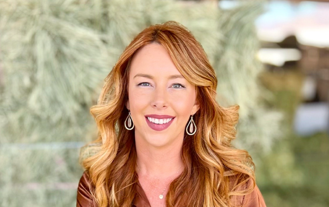 A smiling woman with long strawberry blonde hair sits on hay bales wearing a brown satin blouse.