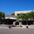 The front of a southwestern-style stucco building with a low wall, arched entry, potted cacti and mesquite trees under a clear sky.
