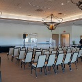 Rows of banquet chairs face a draped table under wrought iron chandeliers in a ballroom, with guests at a catered buffet in the back.