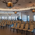 Rows of banquet chairs are set up theater style in a large ballroom with wrought iron chandeliers as guests gather near the windows.