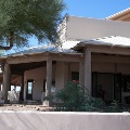 A covered patio with wood posts and a metal roof wraps around a southwestern-style stucco building, with a mesquite tree and barrel cactus in front.