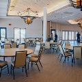 Round banquet tables and chairs fill a ballroom with wrought iron chandeliers as guests mingle near tall windows and glass doors.