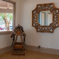 A leather western saddle with a coiled rope rests on a wooden stand beside a window and an ornate gold-framed mirror.