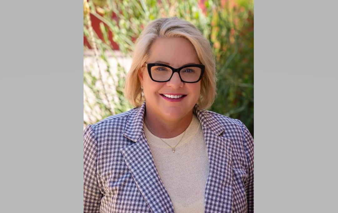 Smiling blonde woman wearing glasses and a checkered blazer outdoors.