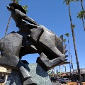 A large angular bronze sculpture of a bucking horse and rider stands among tall palm trees in the Old Town Scottsdale Arts District.