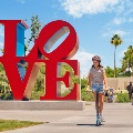 A child wearing a helmet rides a scooter past the large red LOVE sculpture in a park on a sunny day.
