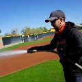 A groundskeeper in a Giants Baseball hoodie waters the infield dirt with a hose at a baseball stadium.