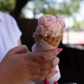 A hand holds a scoop of pink ice cream in a waffle cone wrapped in white paper.