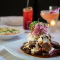 A plated entree topped with pickled red onions and sauce sits on a white tablecloth, with two cocktails and a side dish blurred behind it.