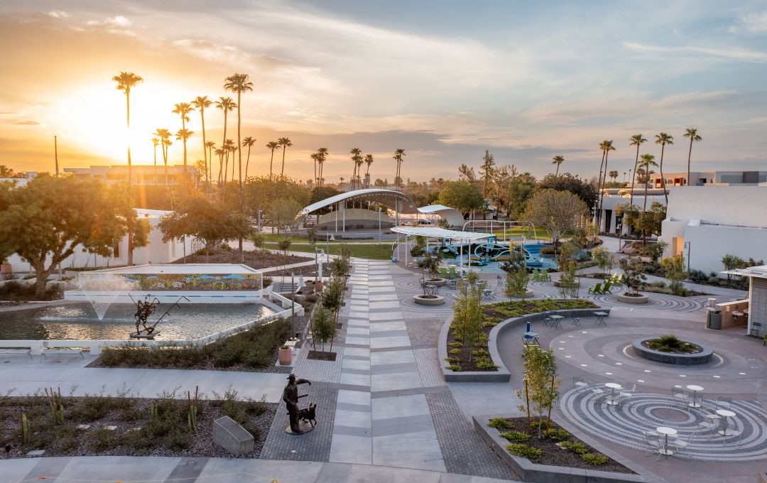 A sunset overlooks a modern plaza with palm trees, a water fountain, sculptures, and a large canopy.