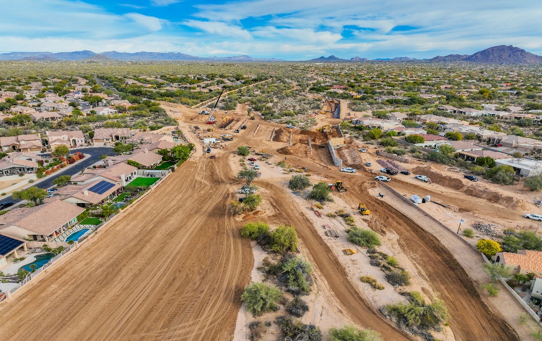 Aerial view of the Rawhide Wash Phase 1 construction site, a wide graded dirt corridor with cranes, excavators, and trucks cutting between two residential neighborhoods, with desert and mountains in the distance.
