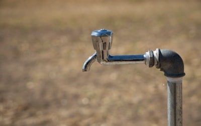 A metal water spigot is attached to a vertical pipe with a blurred brown background.