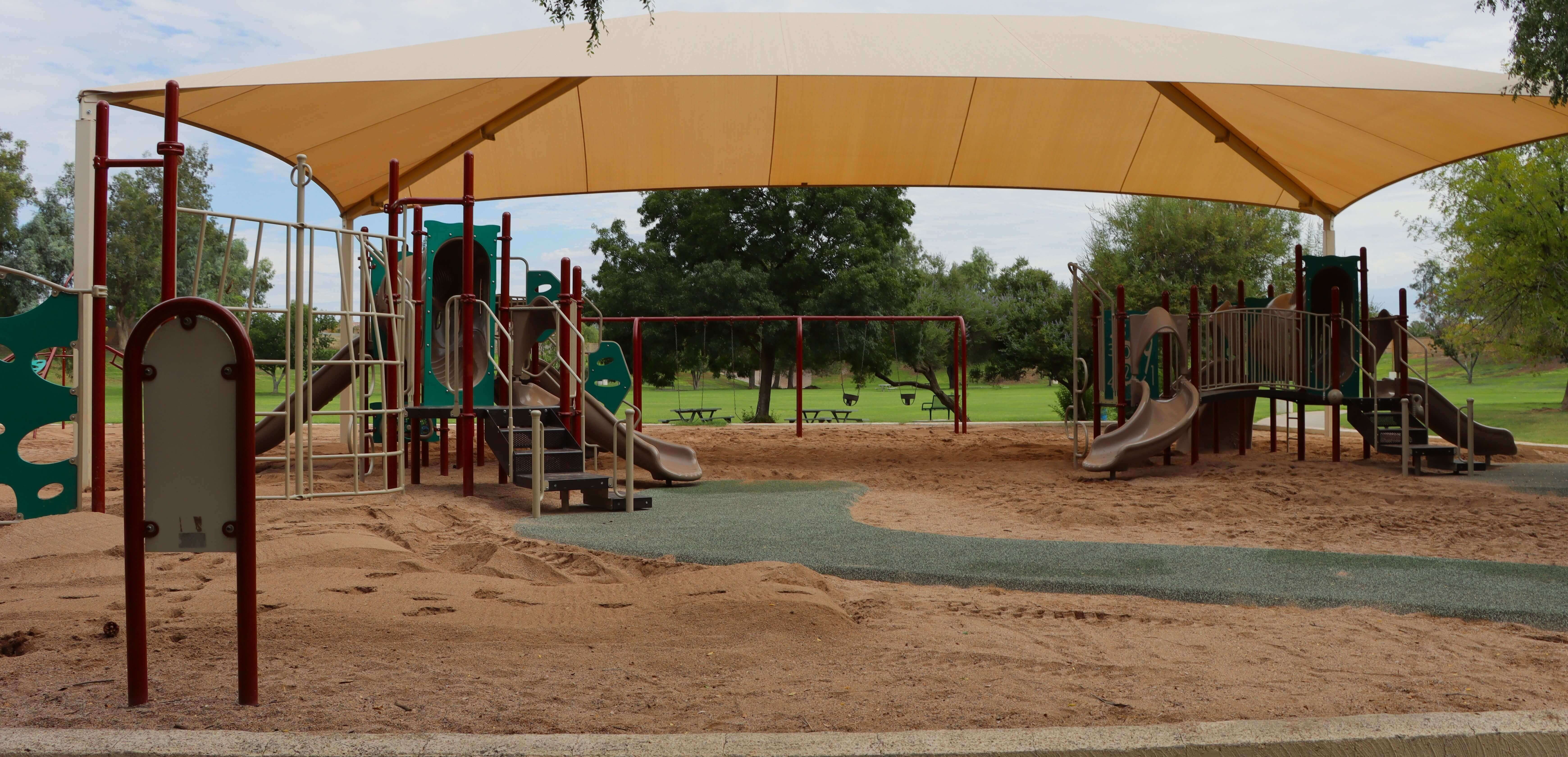 Shaded playground with slides and swings in a sandy park setting.