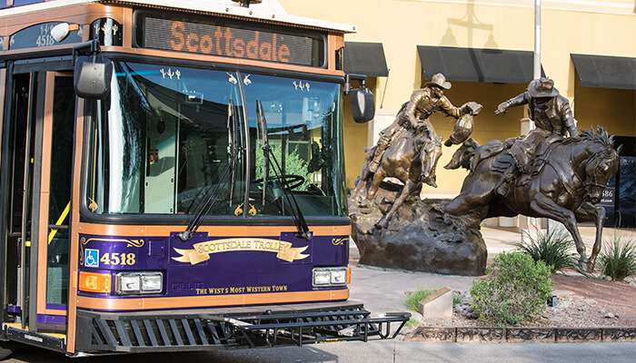Scottsdale trolley passes a bronze cowboy sculpture in a sunny urban streetscape scene.