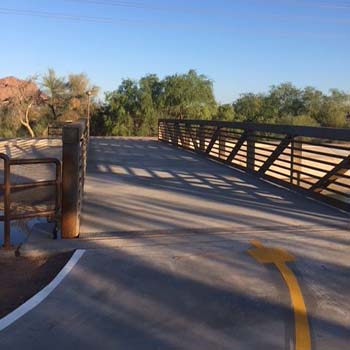 Paved multiuse path crossing a bridge with dark metal railings, desert trees, and directional lane markings under a clear sky.