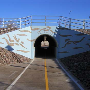 Shared-use path leading into a Scottsdale tunnel beneath a roadway, with painted wall graphics, centerline striping, and metal railings above.