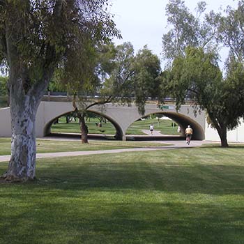 Pedestrians walk through a landscaped park toward a curved concrete underpass bridge surrounded by grass and mature trees.