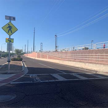 Crosswalk at a roadway underpass with pedestrian warning signs, a beacon signal, sidewalk ramps, and a retaining wall along the right side.