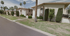 Apartment buildings with traditional lawn landscaping along a residential street, featuring grass, trimmed shrubs, palm trees, and paved walkways under a partly cloudy sky.