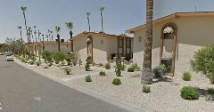 Apartment buildings with sparse desert landscaping along a residential street, featuring gravel ground cover, palm trees, cacti, and small shrubs under a clear sky.