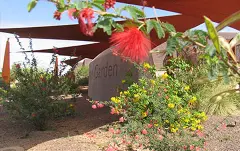 Stone garden sign partially shaded by red shade sails, surrounded by flowering desert plants and gravel, with a red bottlebrush bloom in the foreground.